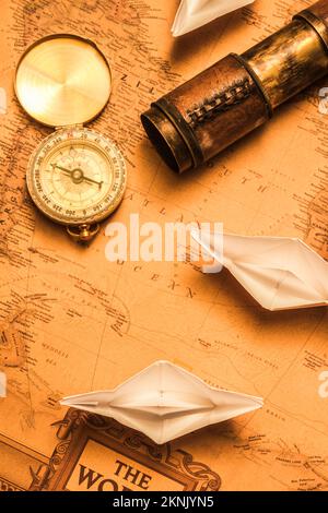 Origami paper boats on a voyage of exploration viewed from above on an ancient chart of the world with vintage brass compass and telescope for navigat Stock Photo