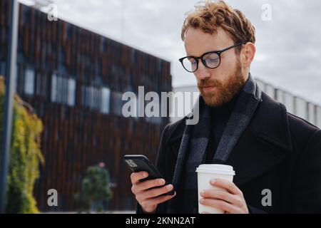 A young red-haired guy, a businessman in glasses, against the backdrop of urban style, checks mail on a smartphone, communicates in a messenger and Stock Photo