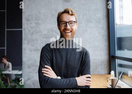 A young red-haired guy, a programmer or an entrepreneur in glasses, in a stylish cafe Looks at the camera and smiles. Freelancer works remotely Stock Photo
