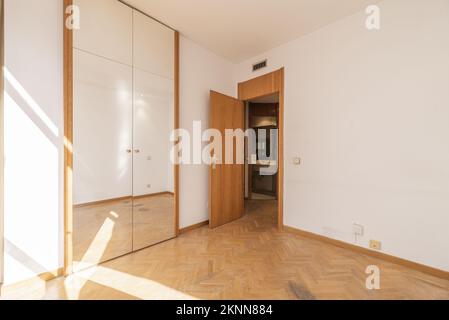 Small room with built-in wardrobe with mirrored doors and sunbeams bouncing off the oak parquet floors Stock Photo