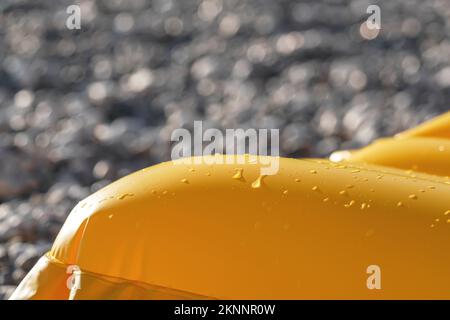 Sun shines to yellow coloured inflatable with drops on water on surface, blurred rocky beach background, closeup detail Stock Photo