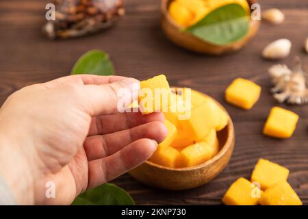 Dried and candied mango cubes in wooden bowls on brown wooden textured background with hand. Side view, close up, selective focus, vegan, vegetarian f Stock Photo