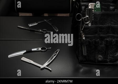 Men's hairdressing tools, hair cutting scissors on a smooth black surface and a black leather bag full of dividers and pockets Stock Photo
