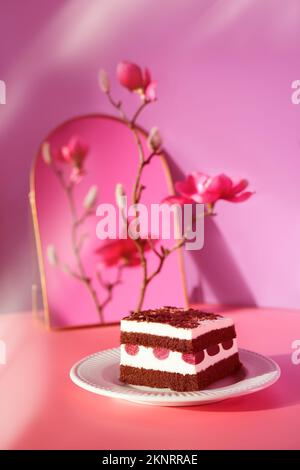Chocolate cake with sour cherries. Piece of cake on a plate with fork. Sweet dessert on pink background with vibrant magenta magnolia blossoms. Stock Photo