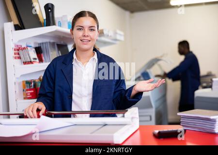 Portrait of cheerful young female typographer in uniform using paper cutter during work in printer shop Stock Photo