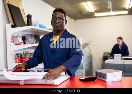 Serious middle-aged African American male in a blue uniform using paper cutter in the printer house Stock Photo