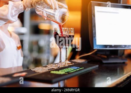 Bartender is pouring red wine into two long-stemmed wineglasses from a measuring cup on the bar counter. Blurred background. Party , cocktail, alcohol Stock Photo