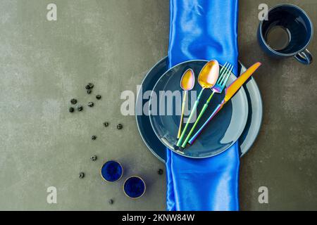 Top view image of various blue porcelain plates with blue tablecloth, gold-rimmed blue glasses, blueberries and iridescent cutlery on off-white backgr Stock Photo