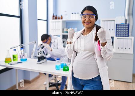 Young hispanic woman expecting a baby working at scientist laboratory screaming proud, celebrating victory and success very excited with raised arms Stock Photo