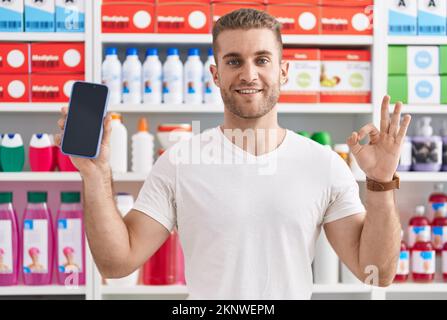 Young caucasian man working at pharmacy drugstore showing smartphone screen doing ok sign with fingers, smiling friendly gesturing excellent symbol Stock Photo