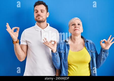 Young brazilian mother and son standing over blue background relaxed and smiling with eyes closed doing meditation gesture with fingers. yoga concept. Stock Photo