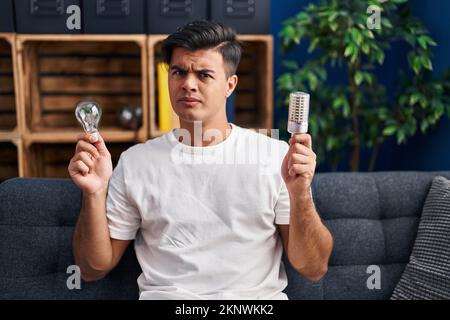 Hispanic man holding led lightbulb and incandescent bulb skeptic and nervous, frowning upset because of problem. negative person. Stock Photo
