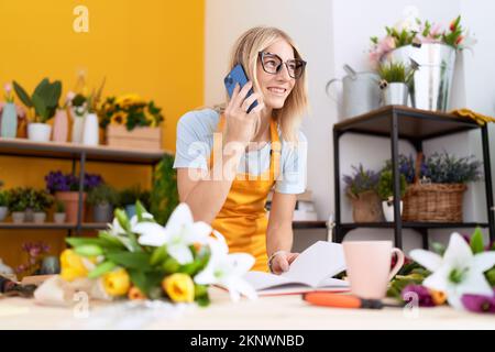 Young blonde woman florist talking on smartphone reading book at flower shop Stock Photo