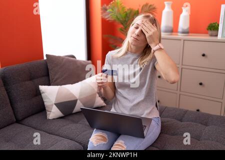 Young blonde woman using laptop and credit card with worried expression at home Stock Photo