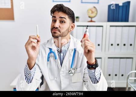 Young doctor man holding electronic cigarette at medical clinic smiling looking to the side and staring away thinking. Stock Photo