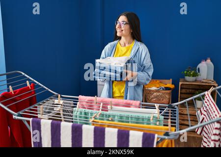 Young hispanic woman holding folded jeans hanging clothes on clothesline at laundry room Stock Photo