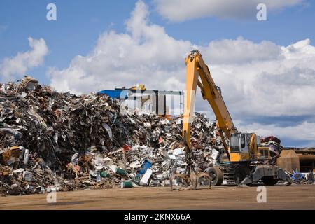 Crane loader fitted with grappling arm in front of pile of discarded metal household and industrial items at scrap metal recycling yard. Stock Photo