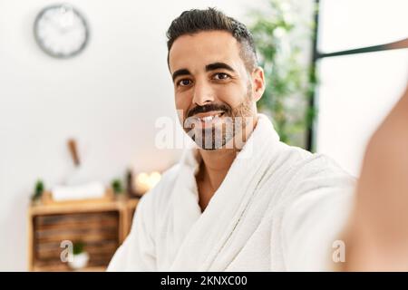 Young hispanic man wearing bathrobe making selfie by the camera at beauty center Stock Photo