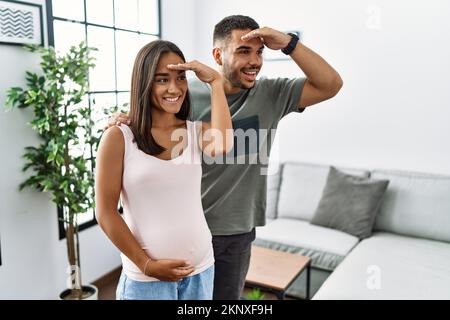 Young interracial couple expecting a baby, touching pregnant belly very happy and smiling looking far away with hand over head. searching concept. Stock Photo