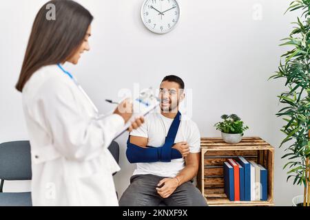 Youg latin injuried man wearing sling talking with the doctor at clinic waiting room. Stock Photo