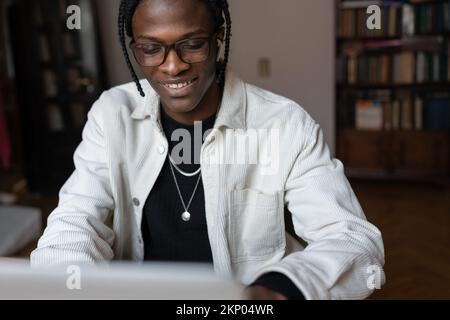 Smiling happy young African man in glasses using laptop at home, spending leisure time on internet Stock Photo