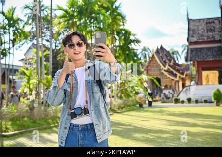 Handsome and happy young Asian male traveler or travel blogger smiling, showing thumb up, taking selfie or video with his smartphone while visiting be Stock Photo