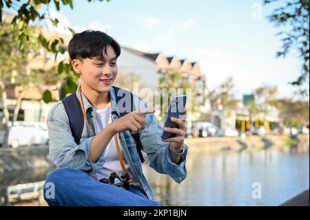 Happy and handsome young Asian male tourist using his smartphone while relaxes sitting by the small canal. Stock Photo