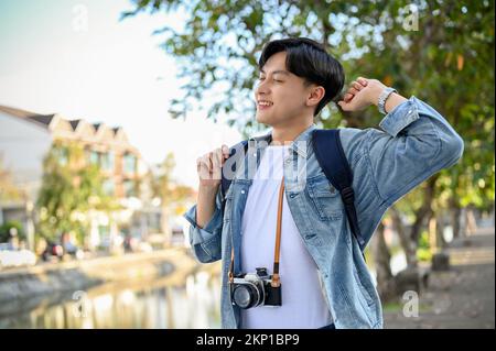 Happy and relaxed young Asian male traveler in jean jacket with his camera strolling around the city, enjoys traveling on his vacation. Stock Photo