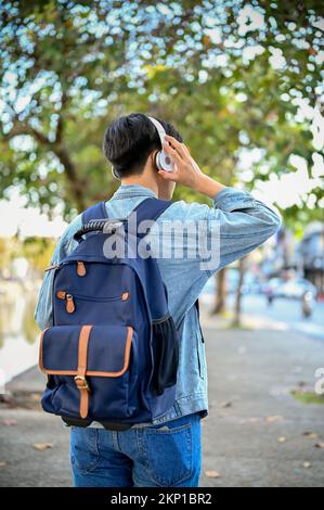 Handsome young Asian male traveler or tourist in jean jacket with his backpack listening to music though his wireless headphones while strolling aroun Stock Photo