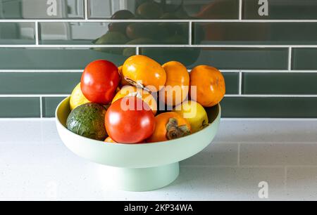 Avocado, tomato, persimmons, and lemons in the green plastic vase on the kitchen countertop Stock Photo