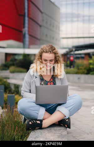 Smiling university student sitting in front of university campus wearing eyeglasses enjoying entertainment content in a laptop. Education internet Stock Photo