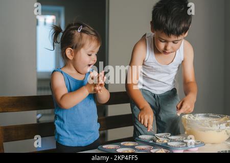Two caucasian little kids making cakes in the kitchen, filling cupcakes. Kids filling muffin forms with raw dough. Sweet food. Stock Photo