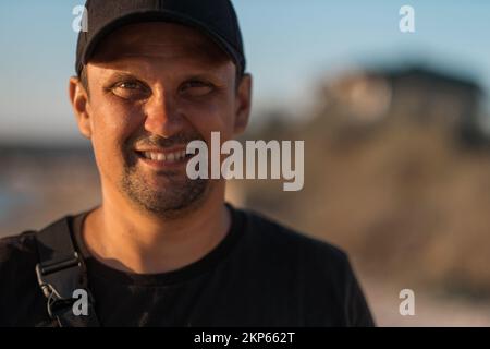 A handsome brunette man in a black t-shirt smiles and looks at the camera against the backdrop of mountains and a blue sky. High quality photo. Stock Photo