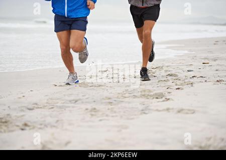 Taking their training seriously. two unrecognizable men jogging together along the beach on an overcast morning. Stock Photo