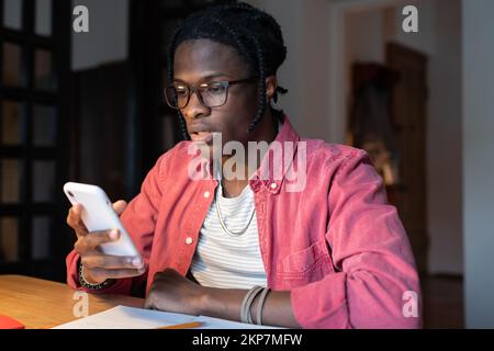 Frustrated African student guy sitting at table holding mobile phone, getting bad news, failed exam Stock Photo