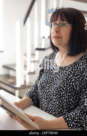 A middle-aged brunette woman in glasses with a bob haircut is reading a book. Stock Photo