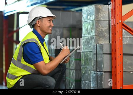 Smiling male warehouse workers checking order details on a tablet while standing near the stack of cardboard boxes Stock Photo