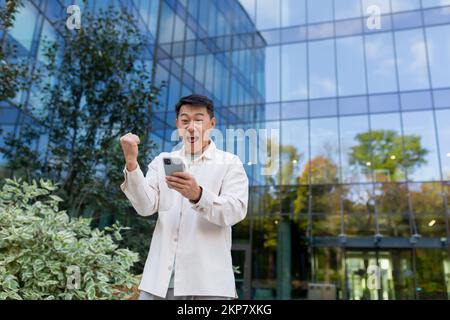Asian businessman in casual clothes with phone outside office building celebrating victory and success, man joyfully holding hand up triumph gesture. Stock Photo