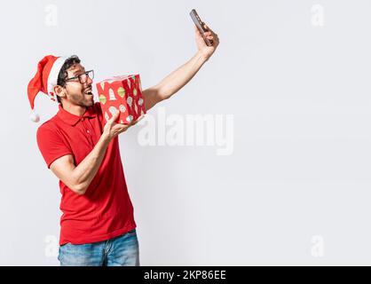 Smiling young man in christmas hat holding a gift and taking a selfie isolated. Christmas handsome man taking a selfie holding a gift isolated. Happy Stock Photo