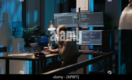 Young adult coder talking on landline phone at night, answering phone call in office. Male programmer using telephone with cord to chat and work on online cloud computing, artificial intelligence. Stock Photo