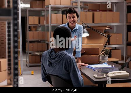 Storehouse workers having fun at job while preparing customers packages putting order in carton box, working in warehouse delivery department. Diverse team checking cargo stock on laptop computer Stock Photo