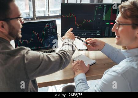 Closeup of two men trader investors sitting at office table together in front of pc, analyzing price flow, discussing rate, actions, monitoring stocks data charts. Selective focus on computer screen Stock Photo