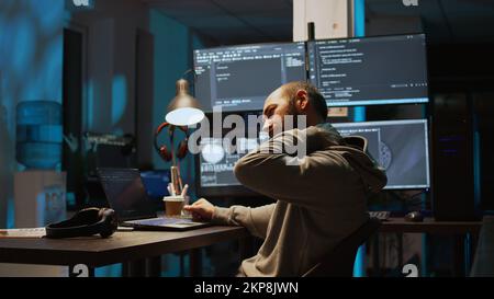 Male web programer writing code and dealing with back pain, sitting at office and feeling spinal discomfort at night. App developer coding html script on terminal window. Handheld shot. Stock Photo