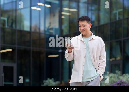 Asian smiling and happy watching online video on phone outside office, man typing message in casual clothes using smartphone. Stock Photo