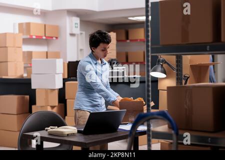 Supervisor putting trendy blouse in cardboard boxes preparing customers orders for delivery, checking shipping detalies on laptop computer in storehouse, Worker analyzing transportation logistics Stock Photo
