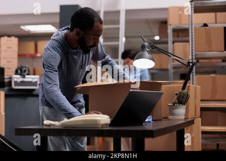 Worker putting client order in cardboard box wrapping with bubble wrap for protection, checking shipping details on laptop computer before delivery package. Employee working in distribution center Stock Photo