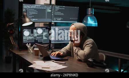 Young programmer talking on phone call in IT developing office, using smartphone for remote conversation. Male engineer working on html code developing, holding mobile phone. Handheld shot. Stock Photo