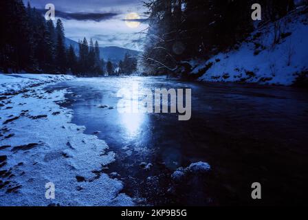 tereblya river in winter in full moon light. snow covered shore with coniferous forest on the shore. mountains beneath a cloudy sky on a frosty night Stock Photo
