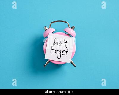 Do not forget reminder notice message. Pink alarm clock with a note paper on blue background with the message do not forget. Stock Photo