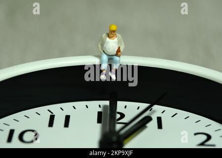 Miniature people toy figure photography. Fat men worker take a break rest above clock. Image photo Stock Photo